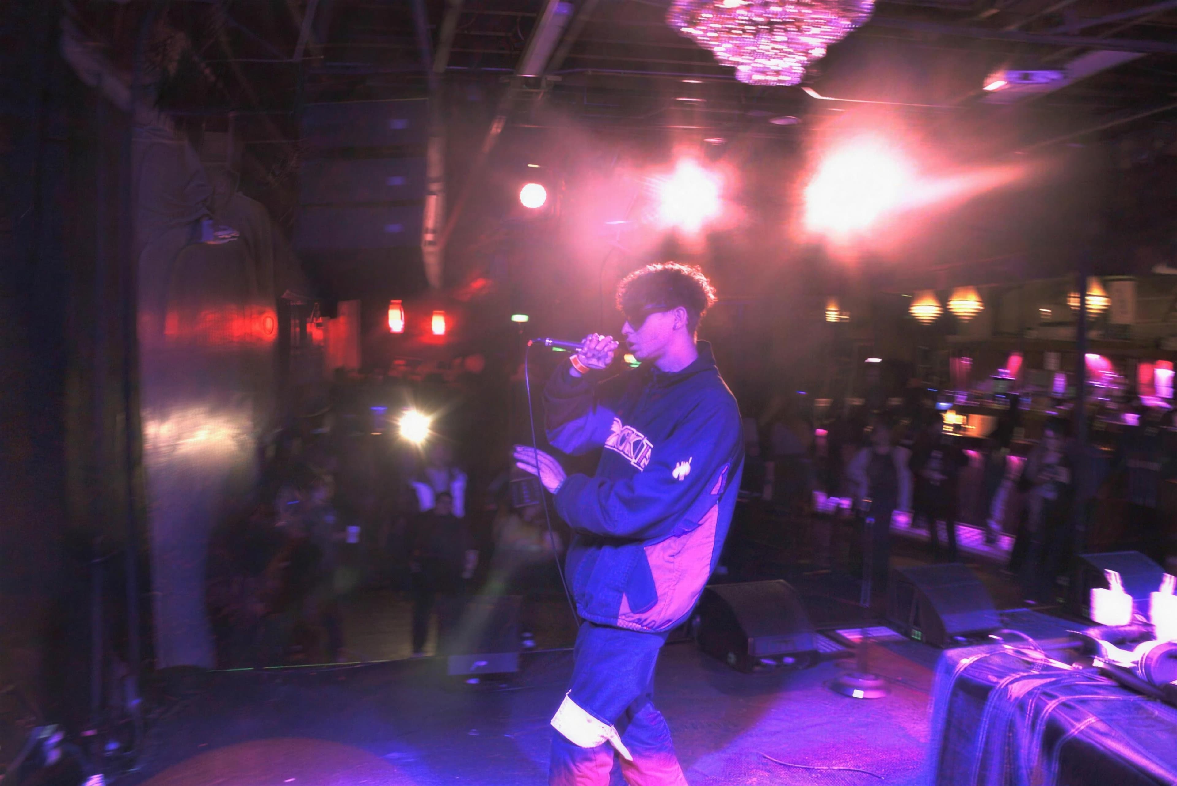 ADRO WHO? on stage, singing into a microphone under violet and pink lights